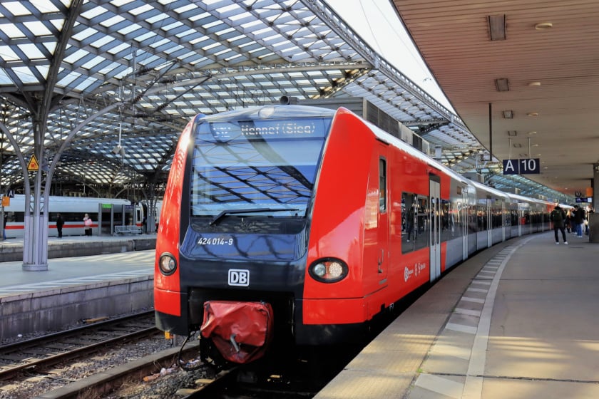 Foto eines roten Regionalzugs der Deutschen Bahn am Bahnhof; auf dem Display steht „Hennef (Sieg)“, rechts Bahnsteigschild „A 10“, Fahrgäste stehen am Gleis unter einer großen Glasdachkonstruktion.