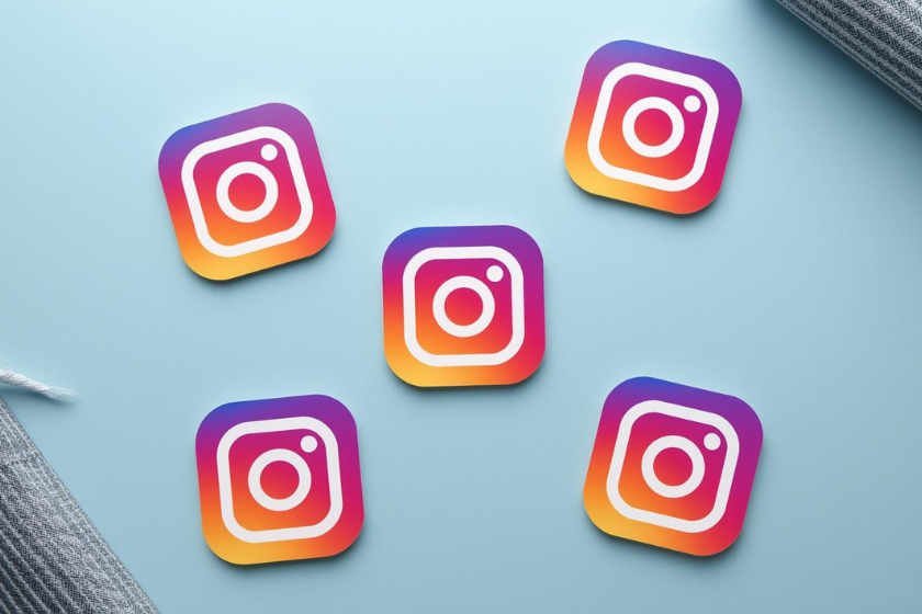 Mehrere Instagram-Logos in bunten Farben auf einem hellblauen Hintergrund, verteilt in einer flachen Anordnung mit dekorativen Stoffelementen am Rand.