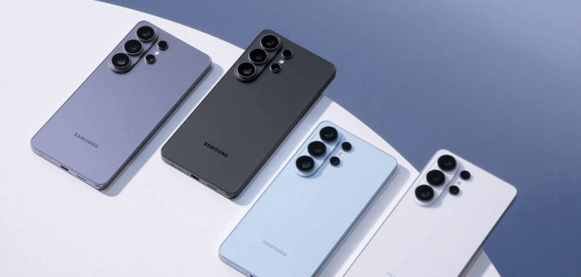Vier Samsung-Smartphones liegen nebeneinander auf einer hellen, runden Oberfläche vor blauem Hintergrund. Die Geräte sind in verschiedenen Farben gehalten (Lila, Schwarz, Hellblau und Weiß) und zeigen jeweils die Rückseite mit mehreren Kameralinsen.