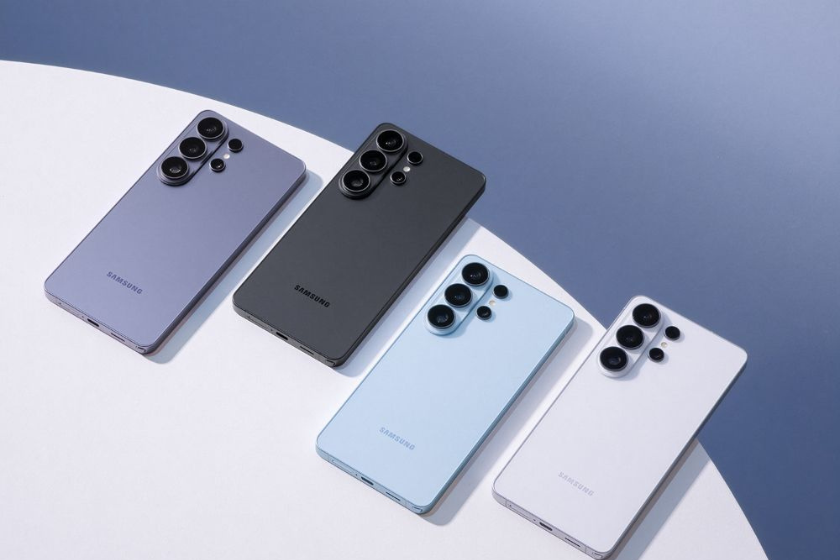 Vier Samsung-Smartphones liegen nebeneinander auf einer hellen, runden Oberfläche vor blauem Hintergrund. Die Geräte sind in verschiedenen Farben gehalten (Lila, Schwarz, Hellblau und Weiß) und zeigen jeweils die Rückseite mit mehreren Kameralinsen.