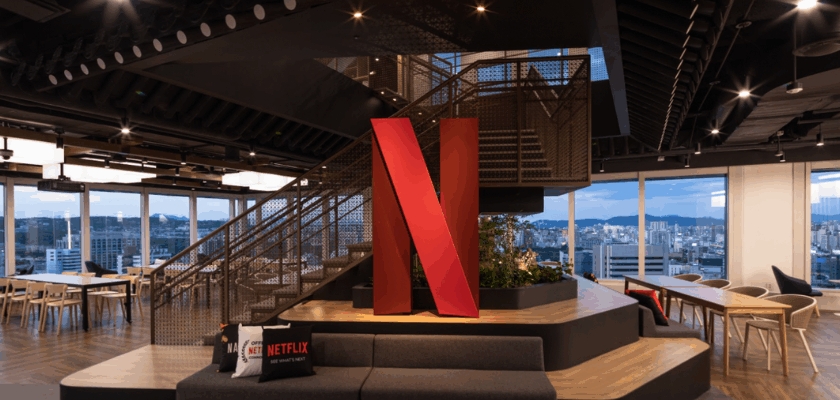 Großes rotes Netflix-Logo in einem modernen Bürogebäude mit Sitzgelegenheiten, Treppe und großen Panoramafenstern mit Blick auf die Stadt.