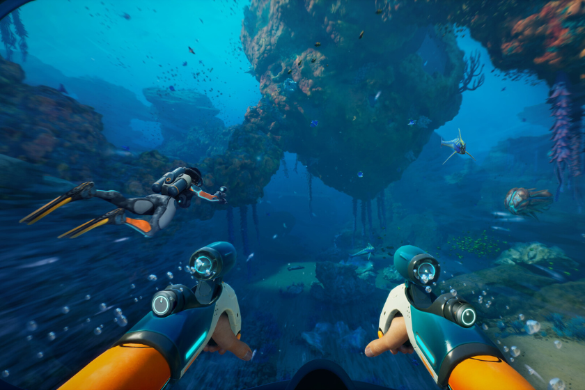 Ego-Perspektive unter Wasser: Eine tauchende Person mit futuristischer Ausrüstung erkundet eine farbenreiche Unterwasserlandschaft mit Felsen, Korallen und verschiedenen Meereslebewesen.