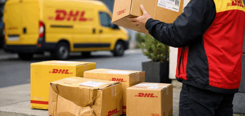Mehrere DHL-Pakete stehen vor einem Wohnhaus, eines davon ist sichtbar beschädigt und eingedrückt. Ein Zusteller in DHL-Kleidung hält ein weiteres Paket in der Hand, während im Hintergrund ein gelber DHL-Transporter zu sehen ist.