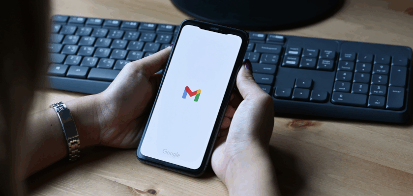 Eine Person hält ein Smartphone in den Händen, auf dessen Display das Gmail-Logo zu sehen ist. Im Hintergrund befinden sich eine schwarze Tastatur und ein Holzschreibtisch, wodurch ein moderner Arbeitsplatz dargestellt wird.