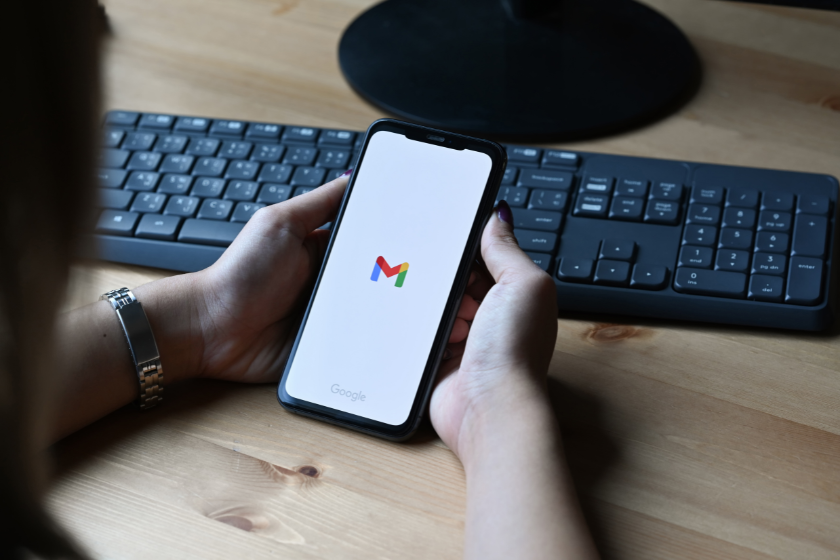 Eine Person hält ein Smartphone in den Händen, auf dessen Display das Gmail-Logo zu sehen ist. Im Hintergrund befinden sich eine schwarze Tastatur und ein Holzschreibtisch, wodurch ein moderner Arbeitsplatz dargestellt wird.