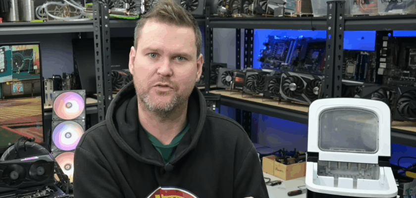 YouTuber TrashBench sitzt in seiner Werkstatt vor mehreren Computerkomponenten und zeigt auf eine Eiswürfelmaschine. Das Gerät wurde in einem Experiment zur Kühlung einer Nvidia GeForce RTX 3060 eingesetzt. Im Hintergrund sind Grafikkarten, Mainboards und ein Testsystem zu sehen.