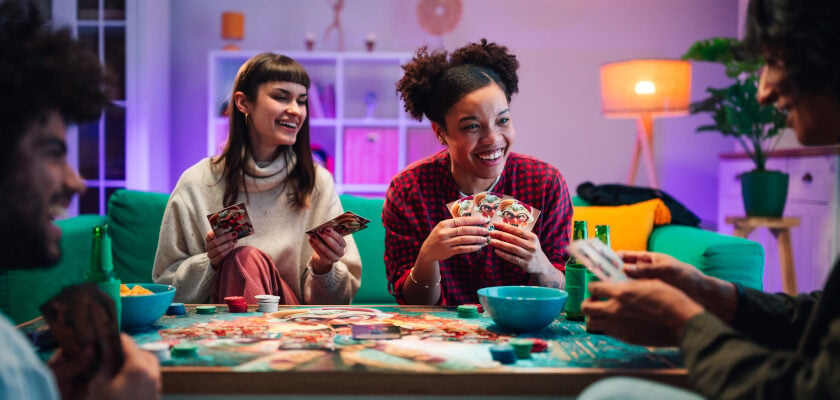 Eine Gruppe von vier Freunden sitzt lachend um einen Tisch und spielt ein Kartenspiel, während im Hintergrund ein gemütliches, warm beleuchtetes Wohnzimmer mit lila und orangefarbenem Licht zu sehen ist. Auf dem Tisch liegen bunte Spielkarten und Chips, dazu stehen Snackschalen und Getränke bereit, während die Spieler ihre Handkarten in die Höhe halten.