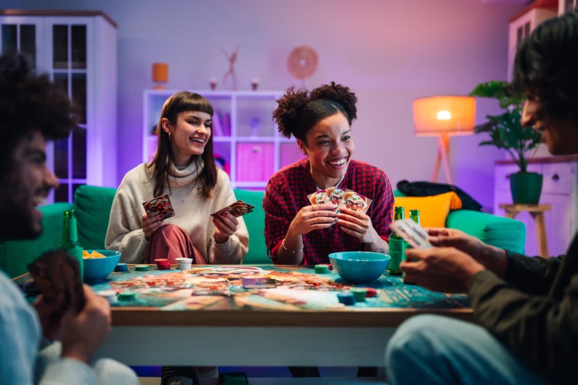 Eine Gruppe von vier Freunden sitzt lachend um einen Tisch und spielt ein Kartenspiel, während im Hintergrund ein gemütliches, warm beleuchtetes Wohnzimmer mit lila und orangefarbenem Licht zu sehen ist. Auf dem Tisch liegen bunte Spielkarten und Chips, dazu stehen Snackschalen und Getränke bereit, während die Spieler ihre Handkarten in die Höhe halten.