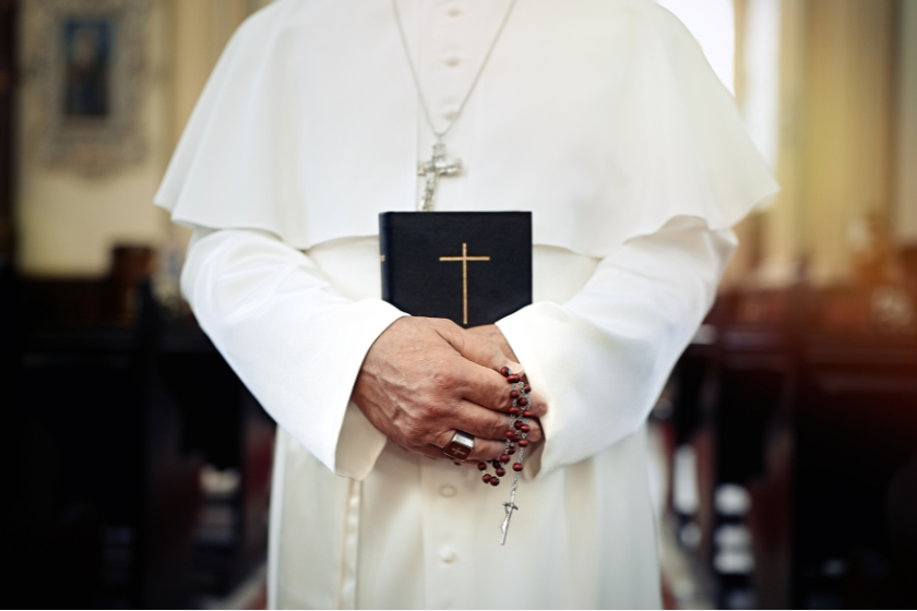 Nahaufnahme einer Person in weißer Papst-Kleidung, die in einer Kirche eine Bibel mit goldenem Kreuz sowie einen Rosenkranz in den Händen hält. Das Bild dient als Symbol für die offizielle Gebets-App des Papstes und die bekannt gewordene Sicherheitslücke.