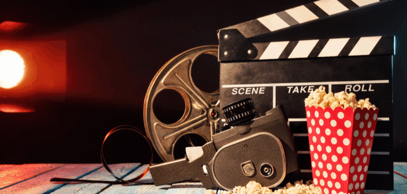 Filmklappe, Filmrolle, Vintage-Filmkamera und Popcorn auf einem Holztisch vor dunklem Hintergrund mit warmem Scheinwerferlicht – symbolisches Motiv für Kinostarts und neue Filme.