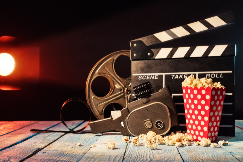 Filmklappe, Filmrolle, Vintage-Filmkamera und Popcorn auf einem Holztisch vor dunklem Hintergrund mit warmem Scheinwerferlicht – symbolisches Motiv für Kinostarts und neue Filme.
