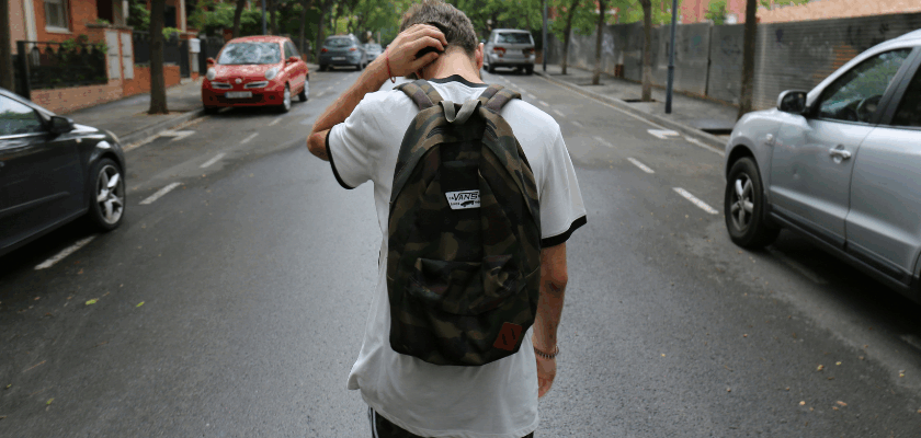 Das Bild zeigt eine Person mit Rucksack, die auf einer ruhigen, von Bäumen gesäumten Straße steht und sich mit einer Hand am Hinterkopf kratzt. Links und rechts der Fahrbahn sind parkende Autos sowie Wohnhäuser zu sehen.