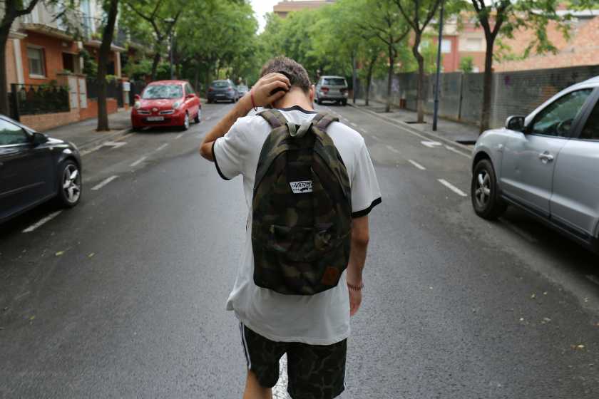 Das Bild zeigt eine Person mit Rucksack, die auf einer ruhigen, von Bäumen gesäumten Straße steht und sich mit einer Hand am Hinterkopf kratzt. Links und rechts der Fahrbahn sind parkende Autos sowie Wohnhäuser zu sehen.
