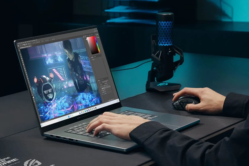 Nahaufnahme zeigt zwei Hände beim Arbeiten an einem ASUS ROG Zephyrus G16 Laptop, auf dessen Bildschirm eine Bildbearbeitungssoftware mit einem futuristischen Cyberpunk-Charakter geöffnet ist. Auf dem dunklen Schreibtisch steht neben dem Laptop ein blau beleuchtetes Streaming-Mikrofon, während eine Hand die Maus bedient und die andere auf der Tastatur ruht.
