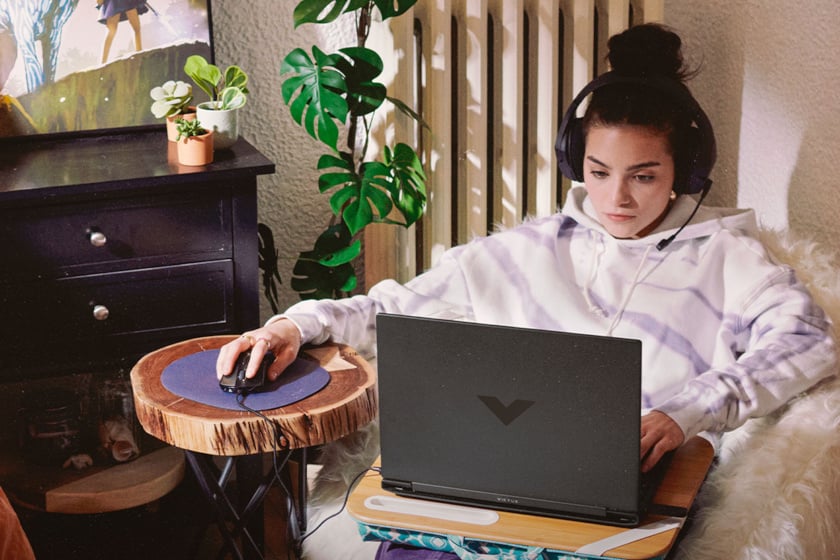 Eine Frau mit hochgestecktem Haar sitzt in einem gemütlichen, pflanzenreichen Zimmer mit Kopfhörern konzentriert vor einem Victus-by-HP-Laptop, der auf einem Bambus-Lapdesk steht. Sie bedient die Maus auf einem rustikalen Holzscheiben-Beistelltisch, während im Hintergrund eine dunkle Kommode mit Topfpflanzen und ein gerahmtes Bild zu sehen sind.
