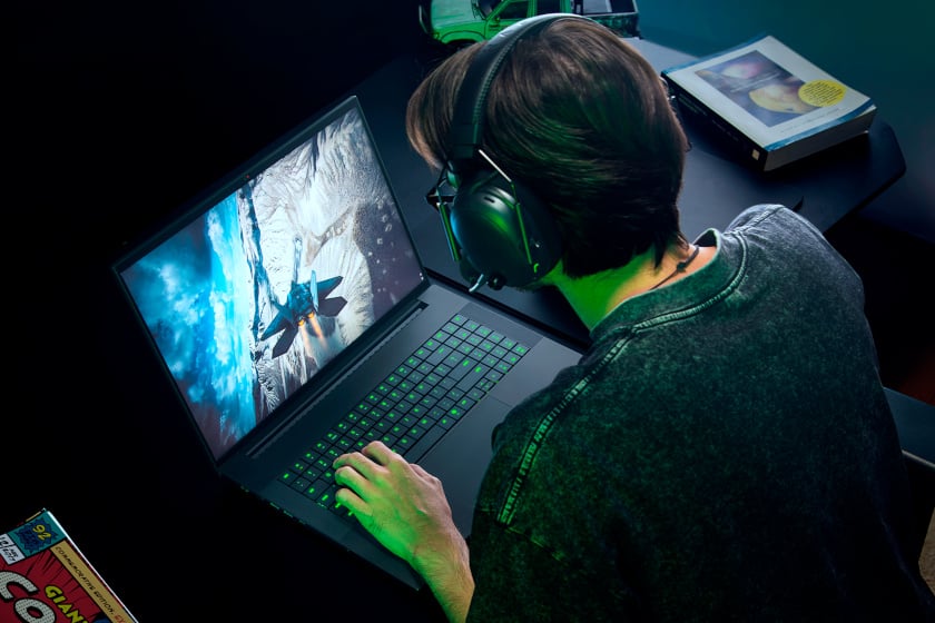 Aufnahme über die Schulter zeigt eine Person mit Kopfhörern, die konzentriert auf einem Razer-Gaming-Laptop mit grün beleuchteter Tastatur spielt. Der Bildschirm zeigt eine winterliche Actionszene mit einem Kampfjet, während im dunklen Raum im Hintergrund ein Buch und ein Comic-Heft auf dem Schreibtisch liegen.