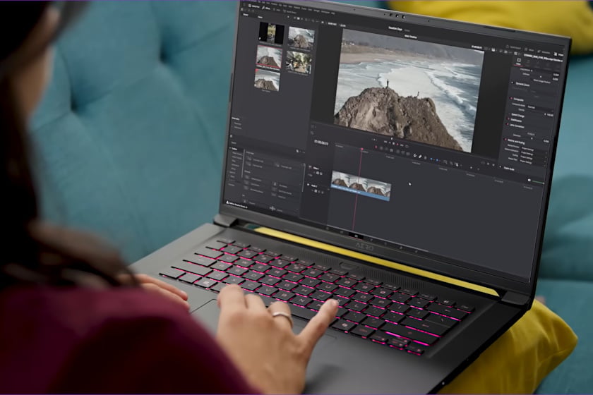 Über-die-Schulter-Aufnahme zeigt eine Person, die auf einem Gigabyte Aero 16 Laptop mit pink beleuchteter Tastatur eine Videobearbeitungssoftware nutzt, in der eine Luftaufnahme einer Küstenlandschaft mit Klippe zu sehen ist. Der Laptop steht auf einer türkisfarbenen Couch, während im Hintergrund gelbe Kissen zu erkennen sind.