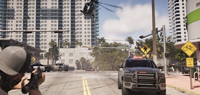 Screenshot aus dem Videospiel „GTA 6“ zeigt eine Verfolgungsjagd-Szene aus der Egoperspektive, bei der ein Charakter mit einem Sturmgewehr auf ein herannahendes Polizeifahrzeug zielt, während ein Polizeihubschrauber tief über die Straße fliegt. Im Hintergrund sind Hochhäuser einer Küstenstadt sowie weitere Polizeifahrzeuge zu erkennen.