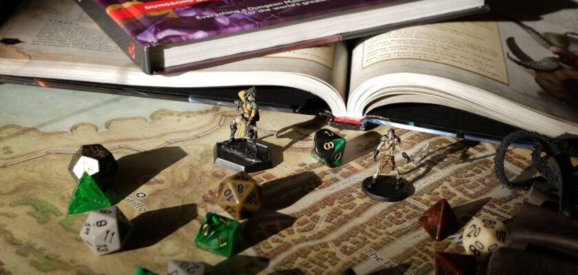 Nahaufnahme eines Dungeons & Dragons-Spieltisches mit einem aufgeschlagenen Regelbuch, das auf einer detaillierten Fantasy-Landkarte liegt. Davor stehen zwei bemalte Miniatur-Figuren, umgeben von zahlreichen mehrseitigen Würfeln in Grün, Schwarz, Weiß und Braun, während im Hintergrund weitere Bücher und Spielutensilien im warmen Licht zu erkennen sind.