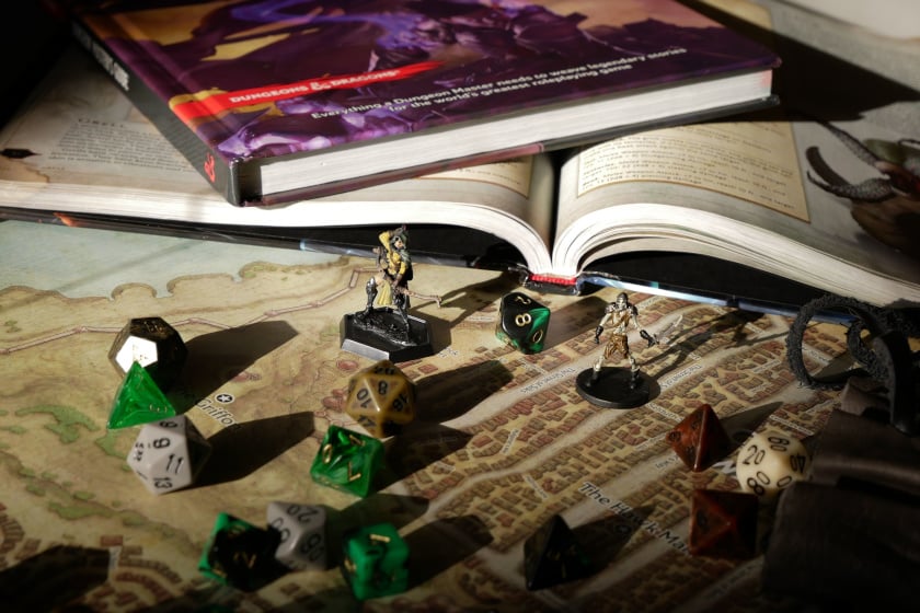 Nahaufnahme eines Dungeons & Dragons-Spieltisches mit einem aufgeschlagenen Regelbuch, das auf einer detaillierten Fantasy-Landkarte liegt. Davor stehen zwei bemalte Miniatur-Figuren, umgeben von zahlreichen mehrseitigen Würfeln in Grün, Schwarz, Weiß und Braun, während im Hintergrund weitere Bücher und Spielutensilien im warmen Licht zu erkennen sind.