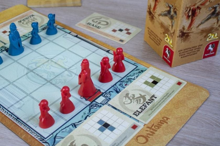 Pegasus Onitama Brettspiel mit roten und blauen Spielfiguren sowie Bewegungskarten auf dem Spielbrett.