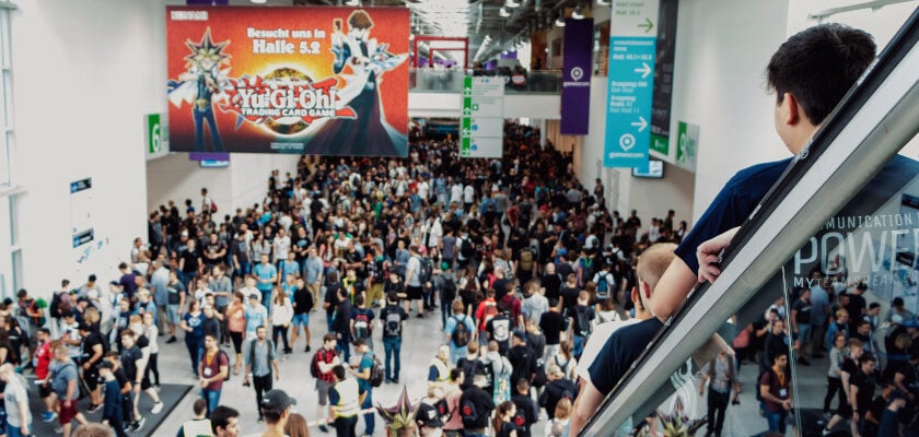 Blick von einer Rolltreppe auf eine dicht gedrängte Besuchermenge in einer Messehalle der gamescom, während im Hintergrund ein großer Bildschirm mit Werbung für „Yu-Gi-Oh! Trading Card Game" hängt. Über der Halle hängen grüne Wegweiser-Schilder von der Decke, und im Vordergrund sind Cosplayer mit auffälligen schwarz-goldenen Kostümaccessoires zu erkennen.