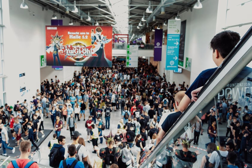Blick von einer Rolltreppe auf eine dicht gedrängte Besuchermenge in einer Messehalle der gamescom, während im Hintergrund ein großer Bildschirm mit Werbung für „Yu-Gi-Oh! Trading Card Game" hängt. Über der Halle hängen grüne Wegweiser-Schilder von der Decke, und im Vordergrund sind Cosplayer mit auffälligen schwarz-goldenen Kostümaccessoires zu erkennen.