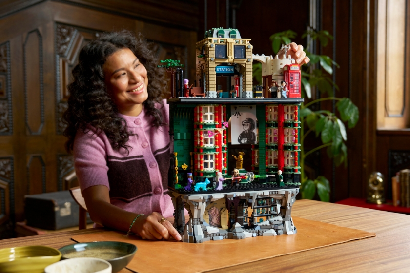 Eine Person präsentiert ein mehrstöckiges LEGO-Modell des Zaubereiministeriums mit offen dargestellten Innenräumen, das auf einem Holztisch in einem Innenraum mit Holzvertäfelung und Pflanzen im Hintergrund steht.