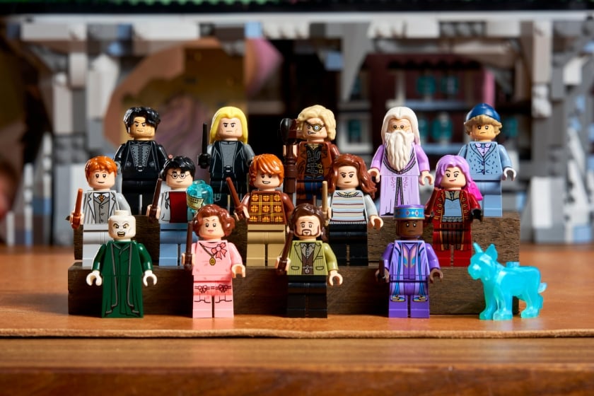 Mehrere LEGO-Minifiguren sowie eine kleine blaue Tierfigur sind in mehreren Reihen auf einer gestuften Präsentationsfläche vor einem unscharfen Hintergrund eines LEGO-Gebäudes angeordnet.