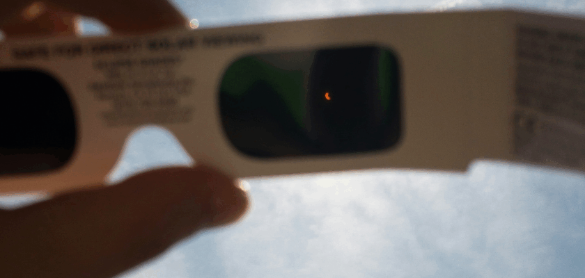 Das Bild zeigt eine Sonnenfinsternis, die durch eine spezielle Schutzbrille beobachtet wird. Die teilweise verdeckte Sonne ist durch das Filterglas als schmale leuchtende Sichel zu erkennen.
