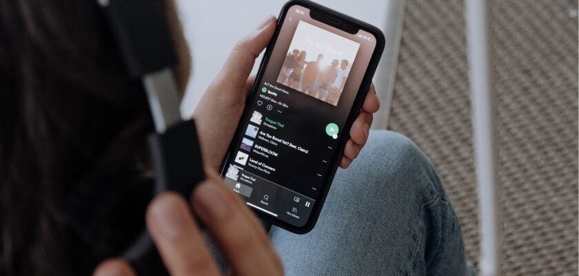 Nahaufnahme über die Schulter einer Person mit Kopfhörern, die ein Smartphone in der Hand hält, auf dem eine Musik-Streaming-App mit Playlist und Songtiteln geöffnet ist. Die Person sitzt entspannt mit angezogenen Knien in Jeans, während im Hintergrund ein weißer Stuhl und ein beiger Teppich zu erkennen sind.