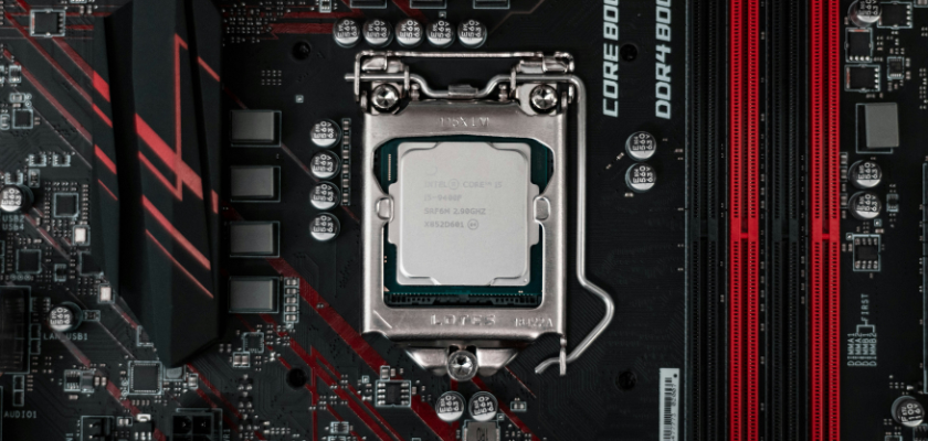 Das Bild zeigt eine Nahaufnahme eines Mainboards mit eingesetztem Intel-Core-i5-Prozessor. Die CPU sitzt mittig im Prozessorsockel und wird von der metallischen Halterung fixiert. Rechts daneben sind mehrere schwarz-rote DDR4-Arbeitsspeicher-Steckplätze zu sehen. Das Mainboard selbst ist überwiegend schwarz und mit roten Designelementen versehen.