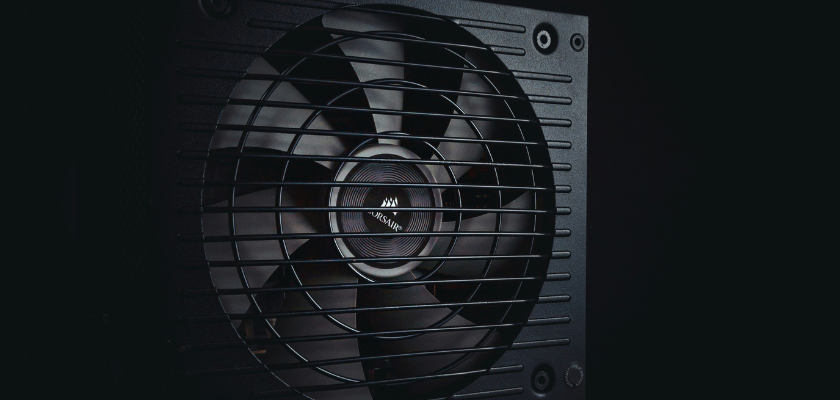 Das Bild zeigt eine Nahaufnahme eines schwarzen PC-Netzteils. Im Mittelpunkt ist der große integrierte Lüfter zu sehen, der von einem schwarzen Schutzgitter abgedeckt wird. Die Aufnahme ist dunkel gehalten und hebt den Lüfter sowie das Gehäuse des Netzteils hervor.