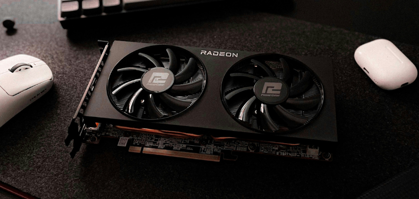 Das Bild zeigt eine schwarze Radeon-Grafikkarte mit zwei großen Lüftern, die auf einer dunklen Schreibtischoberfläche liegt. Links daneben befindet sich eine weiße Computermaus, rechts ein weißes Ladecase für kabellose Kopfhörer. Am oberen Bildrand ist außerdem ein Teil einer Tastatur zu erkennen.