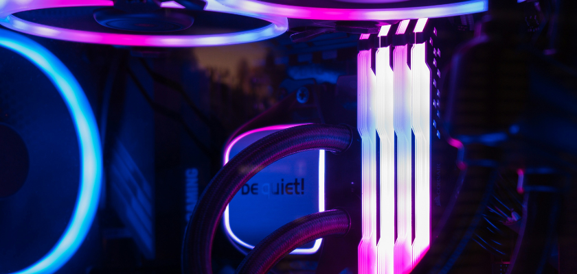 Das Bild zeigt das Innere eines Gaming-PCs mit farbiger RGB-Beleuchtung in Blau-, Pink- und Violetttönen. Im Mittelpunkt sind vier beleuchtete RAM-Module zu sehen. Daneben befinden sich eine Wasserkühlung von be quiet! mit Schläuchen sowie mehrere ebenfalls beleuchtete Lüfter.