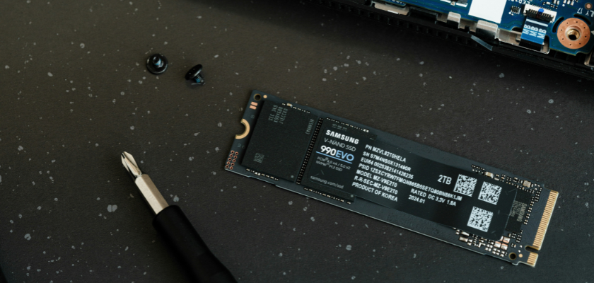 Das Bild zeigt eine Samsung 990 EVO SSD mit 2 TB Speicherkapazität, die auf einer dunklen Arbeitsfläche liegt. Daneben befinden sich ein Schraubendreher und zwei kleine Schrauben. Am oberen Bildrand ist ein Teil einer Platine mit elektronischen Bauteilen zu sehen.