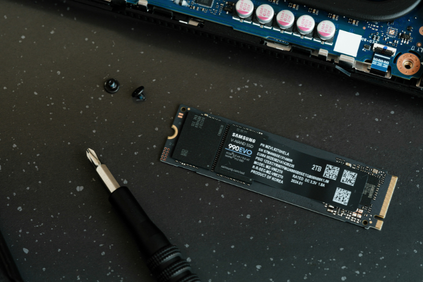 Das Bild zeigt eine Samsung 990 EVO SSD mit 2 TB Speicherkapazität, die auf einer dunklen Arbeitsfläche liegt. Daneben befinden sich ein Schraubendreher und zwei kleine Schrauben. Am oberen Bildrand ist ein Teil einer Platine mit elektronischen Bauteilen zu sehen.
