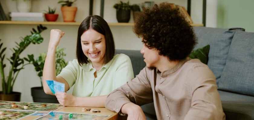 Zwei Personen sitzen gemeinsam im Wohnzimmer und spielen ein Brettspiel. Auf dem Tisch sind Spielpläne, Figuren, Würfel und Karten zu sehen. Die Aufnahme dient als Bebilderung eines Ratgebers zu den besten Brettspielen für Paare.