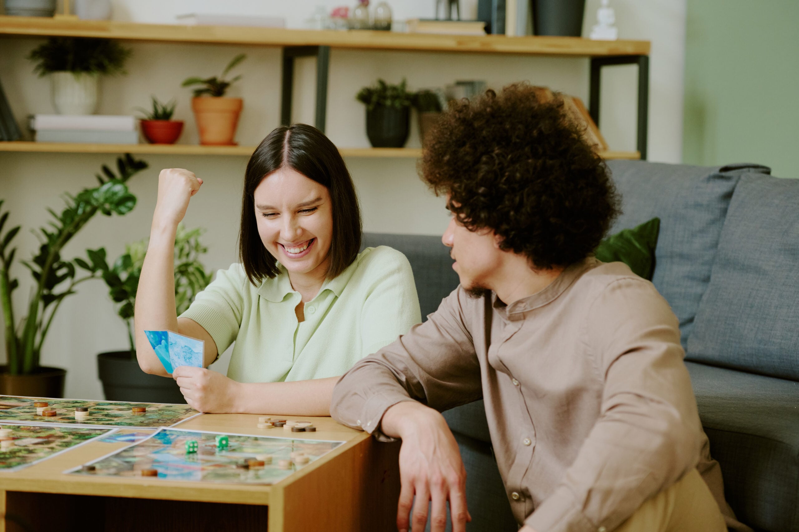 Zwei Personen sitzen gemeinsam im Wohnzimmer und spielen ein Brettspiel. Auf dem Tisch sind Spielpläne, Figuren, Würfel und Karten zu sehen. Die Aufnahme dient als Bebilderung eines Ratgebers zu den besten Brettspielen für Paare.