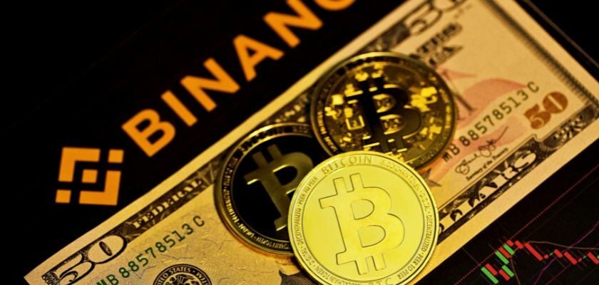 Drei Bitcoin-Münzen liegen auf einem 50-Dollar-Schein. Im Hintergrund sind das Binance-Logo und ein Kursdiagramm zu sehen.