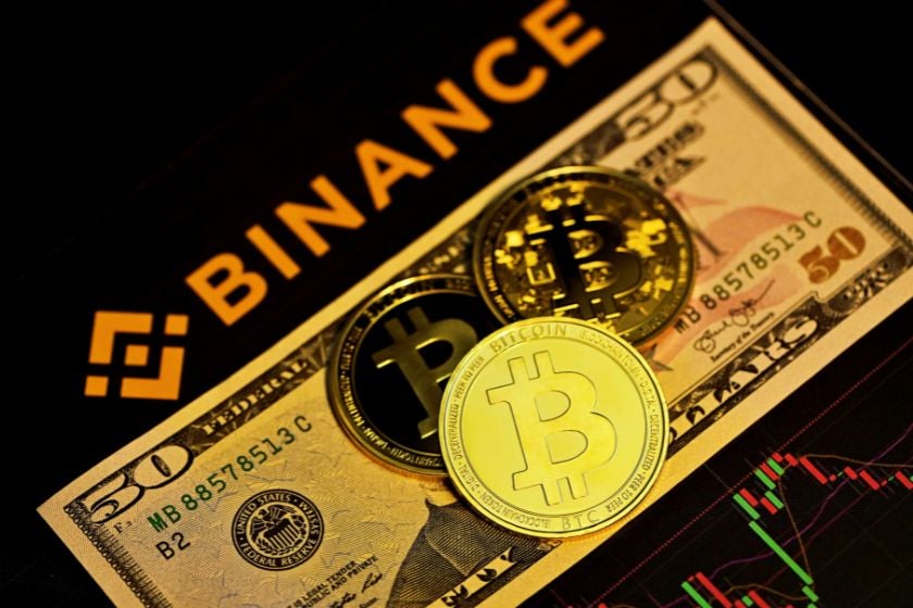 Drei Bitcoin-Münzen liegen auf einem 50-Dollar-Schein. Im Hintergrund sind das Binance-Logo und ein Kursdiagramm zu sehen.