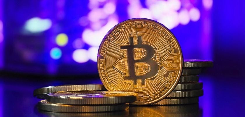 Goldene Bitcoin-Münze mit weiteren gestapelten Münzen vor einem unscharfen, violett beleuchteten Hintergrund.