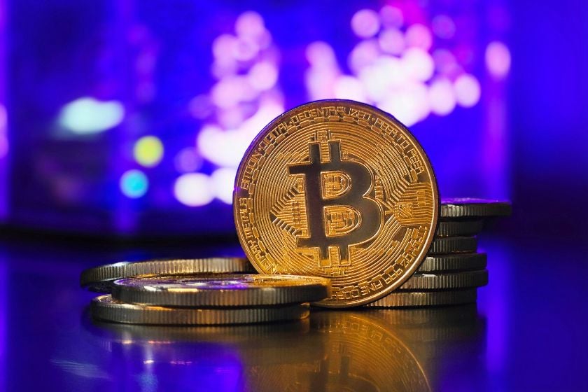 Goldene Bitcoin-Münze mit weiteren gestapelten Münzen vor einem unscharfen, violett beleuchteten Hintergrund.